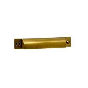 May include: A brass drawer pull with rounded edges and a subtle brushed finish. The pull has two visible screws on either end.