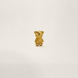 May include: A small gold-coloured teddy bear charm. The bear is sitting with its arms and legs outstretched.