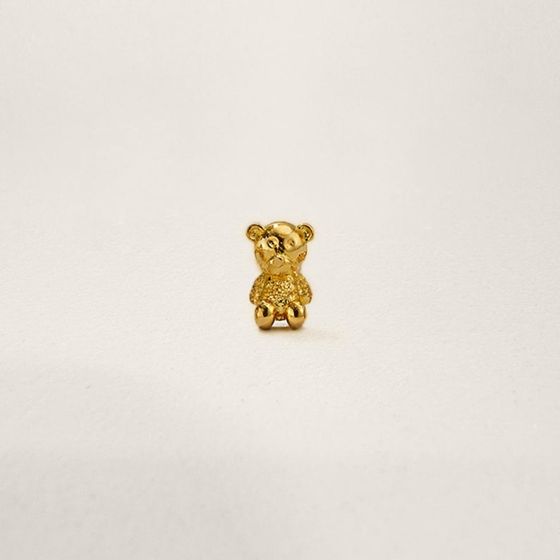 May include: A small gold-coloured teddy bear charm. The bear is sitting with its arms and legs outstretched.