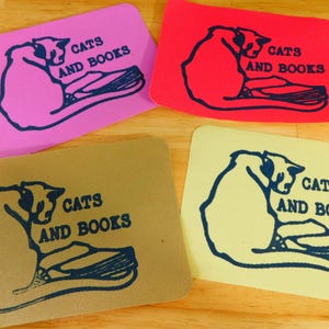 May include: Four rectangular coasters in pink, red, tan, and yellow, each featuring a black illustration of a cat and books, with the words "CATS AND BOOKS" in black text. The coasters are arranged on a wooden surface.