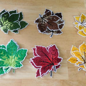May include: Six embroidered leaf patches in various colors: green, brown, yellow, and red. Each leaf has a white border and detailed veining. These decorative patches are ideal for adding to clothing or accessories.