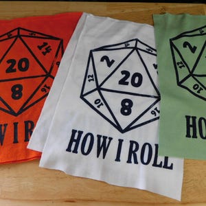 May include: Three fabric squares in orange, white, and green, each featuring a black geometric design with the numbers 2, 8, 10, 12, 14, 16, and 20. The text "HOW I ROLL" is printed below the design.