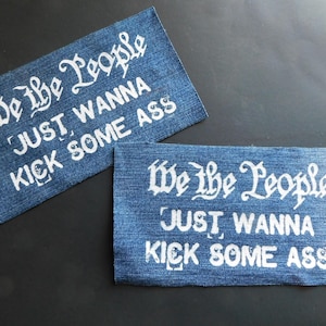 May include: Two denim patches with white text. The patches read "We the People JUST WANNA KICK SOME ASS" in a stylized font. The patches are rectangular and have a frayed edge.