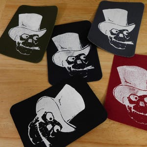 May include: A collection of rectangular patches featuring a skull wearing a top hat and smoking a cigar. The patches are in various colors, including black, red, gray, and olive green, with a white graphic.