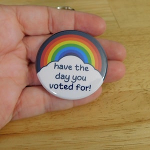 May include: A round button with a rainbow design and the text "have the day you voted for!". The rainbow arcs over a white cloud, set against a dark blue border. The button is held in a hand.