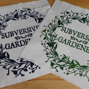 May include: Two white fabric squares with the words "SUBVERSIVE GARDENER" in dark blue and green, surrounded by a floral wreath design. The fabric has a slightly textured appearance.