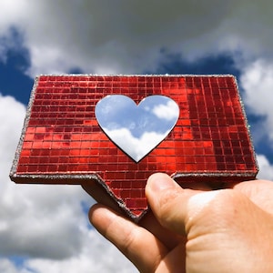 May include: A red, sparkly, heart-shaped like button with a silver heart in the centre reflecting the sky and clouds. The button is being held up in the air.