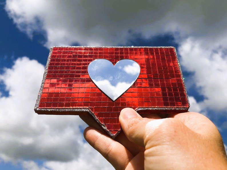 May include: A red, sparkly, heart-shaped like button with a silver heart in the centre reflecting the sky and clouds. The button is being held up in the air.