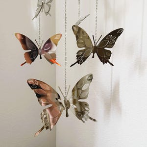 May include: Several metal butterfly ornaments hang from silver chains. The butterflies have intricate wing designs and are colored in shades of brown, copper, and silver. The butterflies are suspended in front of a white wall.