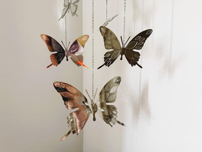 May include: Several metal butterfly ornaments hang from silver chains. The butterflies have intricate wing designs and are colored in shades of brown, copper, and silver. The butterflies are suspended in front of a white wall.