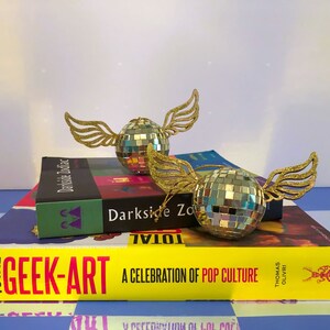 Puede incluir: Dos adornos decorativos de bola de discoteca con alas doradas brillantes descansan sobre una pila de libros. Los libros incluyen t&iacute;tulos como "Darkside Zodiac" y "Total Geek-Art: A Celebration of Pop Culture". El fondo tiene un patr&oacute;n de cuadros azules y blancos.