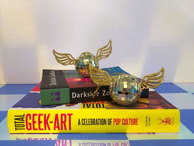 Puede incluir: Dos adornos decorativos de bola de discoteca con alas doradas brillantes descansan sobre una pila de libros. Los libros incluyen t&iacute;tulos como "Darkside Zodiac" y "Total Geek-Art: A Celebration of Pop Culture". El fondo tiene un patr&oacute;n de cuadros azules y blancos.