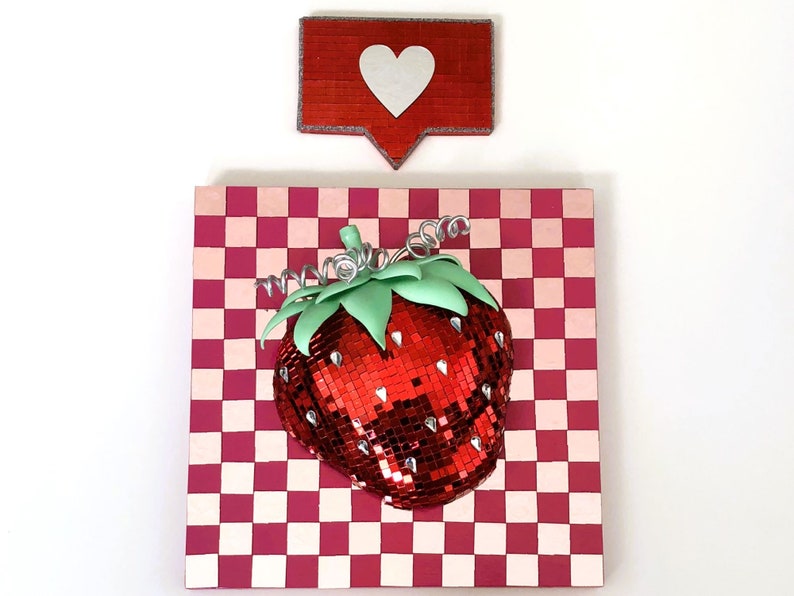 May include: A red and pink checkered background with a red, sparkly strawberry on top. A red heart icon with a white outline is above the strawberry.