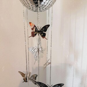 May include: A decorative mobile featuring a mirrored disco ball at the top, with several butterfly ornaments hanging below. The butterflies are in various colors, including black, silver, and brown, and are suspended by silver chains.