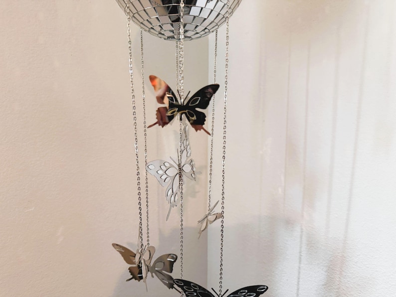 May include: A decorative mobile featuring a mirrored disco ball at the top, with several butterfly ornaments hanging below. The butterflies are in various colors, including black, silver, and brown, and are suspended by silver chains.
