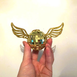Puede incluir: Una bola de discoteca dorada con alas brillantes. La bola est&aacute; hecha de peque&ntilde;os cuadrados reflectantes, y las alas est&aacute;n cortadas de papel de purpurina dorada. El objeto se sostiene en una mano sobre un fondo blanco.