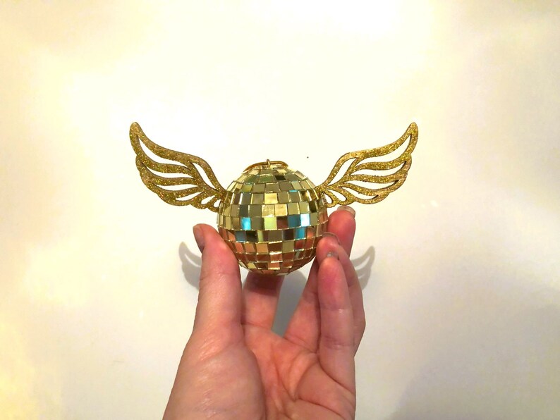 Puede incluir: Una bola de discoteca dorada con alas brillantes. La bola est&aacute; hecha de peque&ntilde;os cuadrados reflectantes, y las alas est&aacute;n cortadas de papel de purpurina dorada. El objeto se sostiene en una mano sobre un fondo blanco.