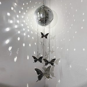 May include: A silver disco ball hangs from the ceiling, reflecting light onto the white wall. Below the disco ball, several black butterfly ornaments are suspended by chains, creating a whimsical and decorative display.