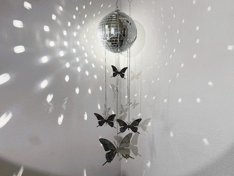 May include: A silver disco ball hangs from the ceiling, reflecting light onto the white wall. Below the disco ball, several black butterfly ornaments are suspended by chains, creating a whimsical and decorative display.