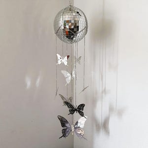 May include: A silver disco ball mobile with hanging butterfly ornaments. The butterflies are in various shades of silver and black, suspended by delicate chains. The mobile is hanging in front of a white wall.