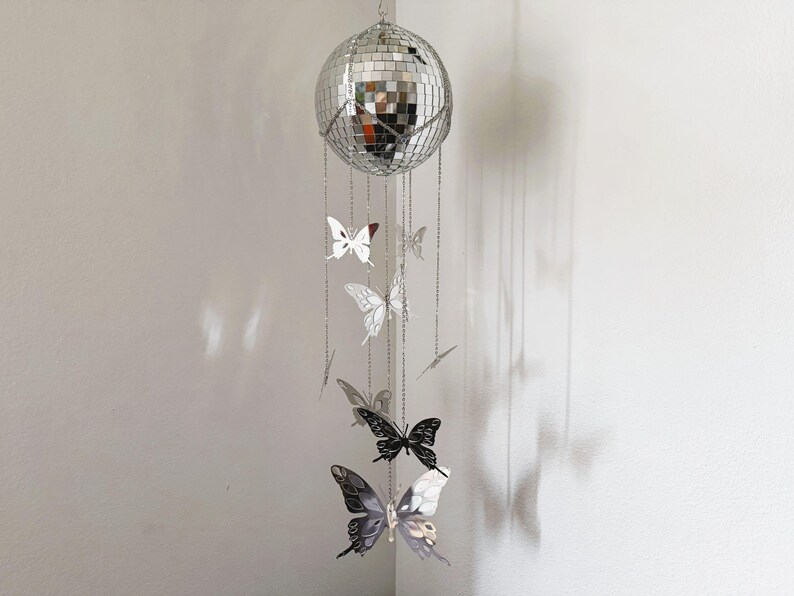 May include: A silver disco ball mobile with hanging butterfly ornaments. The butterflies are in various shades of silver and black, suspended by delicate chains. The mobile is hanging in front of a white wall.