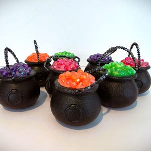 May include: A collection of seven small black plastic cauldrons, each featuring a dark beaded handle and filled with vibrant, multi-colored translucent beads in shades of orange, green, purple, and pink, arranged in a cluster on a white surface.