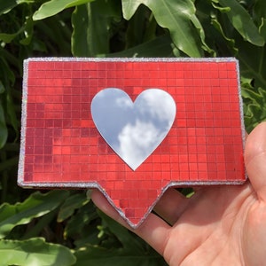May include: A red and silver glitter heart-shaped icon with a mirrored surface. The icon is shaped like the Instagram like button.