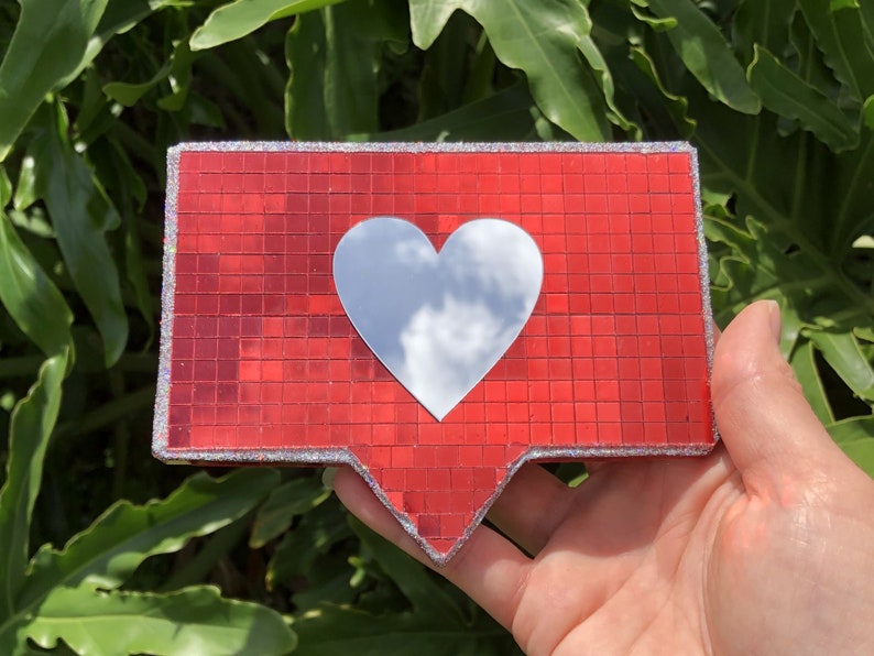 May include: A red and silver glitter heart-shaped icon with a mirrored surface. The icon is shaped like the Instagram like button.