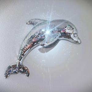 Peut inclure: Une sculpture de dauphin en mosaïque argentée avec une surface réfléchissante. Le dauphin est monté sur un mur blanc, et les carreaux de mosaïque créent un effet scintillant. L'objet est décoratif.