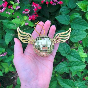 Puede incluir: Adorno de bola de discoteca dorada con alas doradas adjuntas. El adorno es redondo y est&aacute; cubierto de peque&ntilde;os cuadrados reflectantes. Las alas est&aacute;n recortadas con un dise&ntilde;o detallado de plumas. Se adjunta una cuerda dorada para colgar. Fondo de jard&iacute;n verde exuberante.