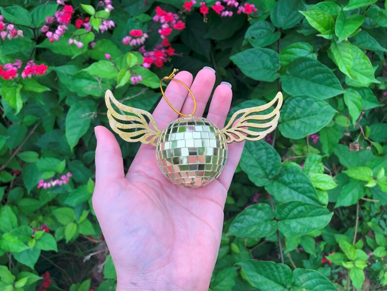 Puede incluir: Adorno de bola de discoteca dorada con alas doradas adjuntas. El adorno es redondo y est&aacute; cubierto de peque&ntilde;os cuadrados reflectantes. Las alas est&aacute;n recortadas con un dise&ntilde;o detallado de plumas. Se adjunta una cuerda dorada para colgar. Fondo de jard&iacute;n verde exuberante.