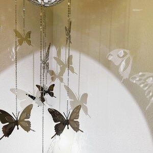 May include: A decorative mobile featuring butterfly cutouts in varying sizes and shades of brown and silver. The butterflies are suspended from a mirrored sphere by delicate chains. Shadows of the butterflies are cast on the wall.