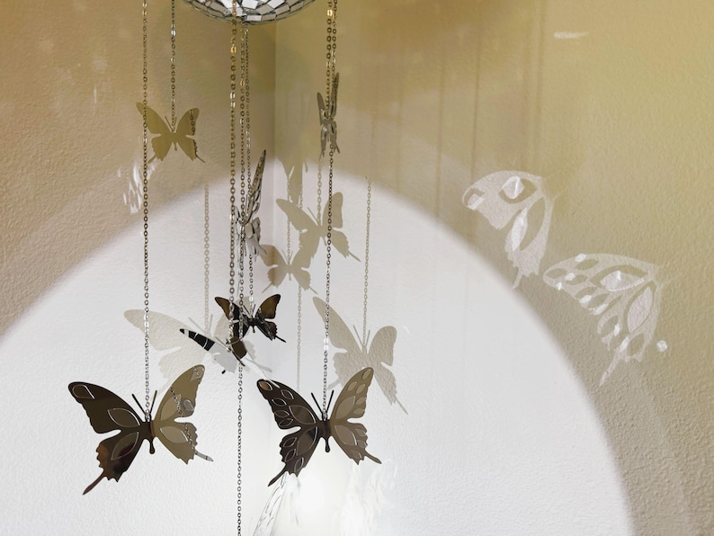 May include: A decorative mobile featuring butterfly cutouts in varying sizes and shades of brown and silver. The butterflies are suspended from a mirrored sphere by delicate chains. Shadows of the butterflies are cast on the wall.
