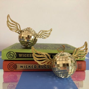 Puede incluir: Dos adornos dorados de bola de discoteca con alas brillantes descansan sobre dos libros. Los libros son verdes y naranjas con las palabras "Wicked Bugs" en los lomos. Los adornos son reflectantes y esf&eacute;ricos.