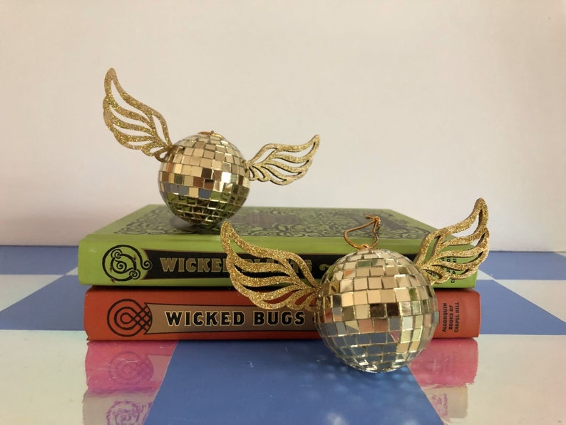 Puede incluir: Dos adornos dorados de bola de discoteca con alas brillantes descansan sobre dos libros. Los libros son verdes y naranjas con las palabras "Wicked Bugs" en los lomos. Los adornos son reflectantes y esf&eacute;ricos.