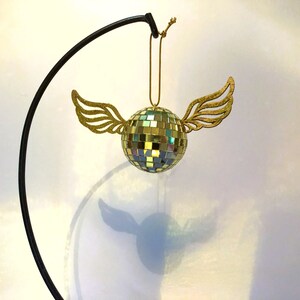 Puede incluir: Adorno decorativo de bola de discoteca con alas doradas brillantes. La bola est&aacute; hecha de peque&ntilde;os cuadrados reflectantes y cuelga de una cuerda dorada. El adorno est&aacute; suspendido de un soporte de metal negro.