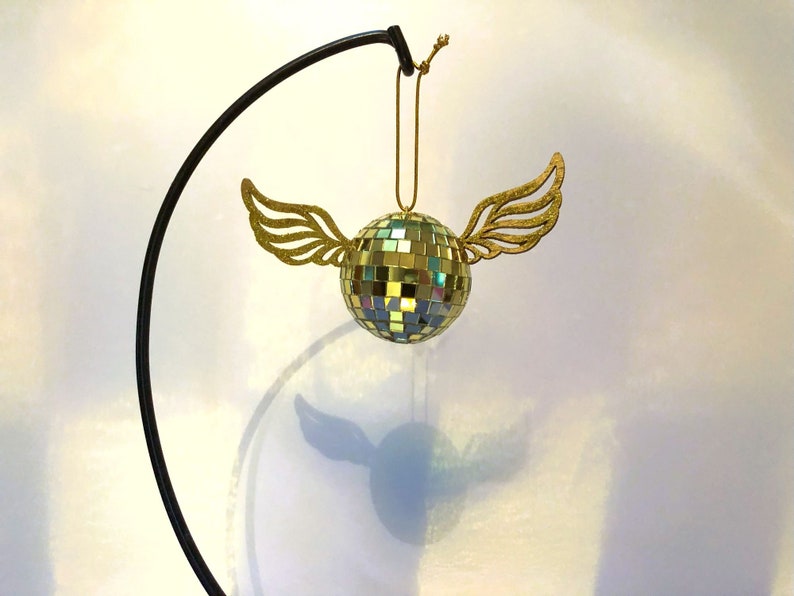 Puede incluir: Adorno decorativo de bola de discoteca con alas doradas brillantes. La bola est&aacute; hecha de peque&ntilde;os cuadrados reflectantes y cuelga de una cuerda dorada. El adorno est&aacute; suspendido de un soporte de metal negro.