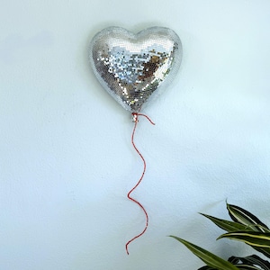 May include: A heart-shaped decoration made of small, reflective silver tiles, resembling a disco ball. It hangs from a red string against a white wall, with green and white leafy plants in the lower right corner.