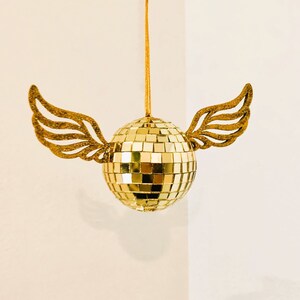 Puede incluir: Adorno dorado de bola de discoteca con alas brillantes adjuntas. La bola est&aacute; hecha de peque&ntilde;os cuadrados reflectantes y cuelga de un hilo dorado. Las alas doradas a&ntilde;aden un toque caprichoso.