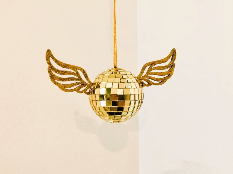 Puede incluir: Adorno dorado de bola de discoteca con alas brillantes adjuntas. La bola est&aacute; hecha de peque&ntilde;os cuadrados reflectantes y cuelga de un hilo dorado. Las alas doradas a&ntilde;aden un toque caprichoso.