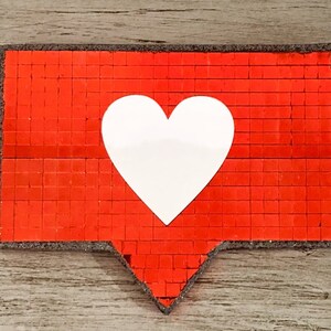 May include: A red heart-shaped button with a white heart in the centre. The button is made of small, square sequins.
