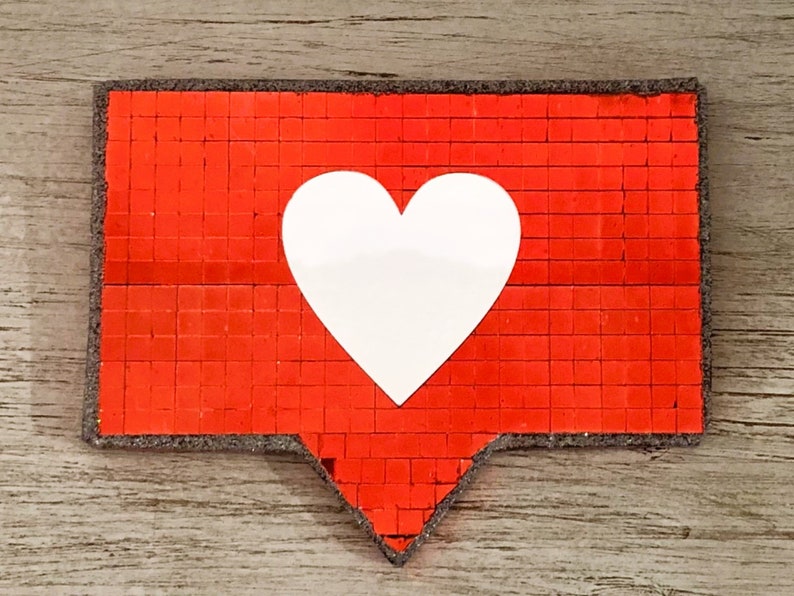 May include: A red heart-shaped button with a white heart in the centre. The button is made of small, square sequins.