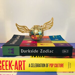 Puede incluir: Una bola de discoteca dorada con alas brillantes descansa sobre dos libros. El libro superior se titula "Darkside Zodiac" y el libro inferior se titula "Total Geek-Art: A Celebration of Pop Culture". Los libros est&aacute;n apilados sobre una superficie a rayas moradas y blancas.