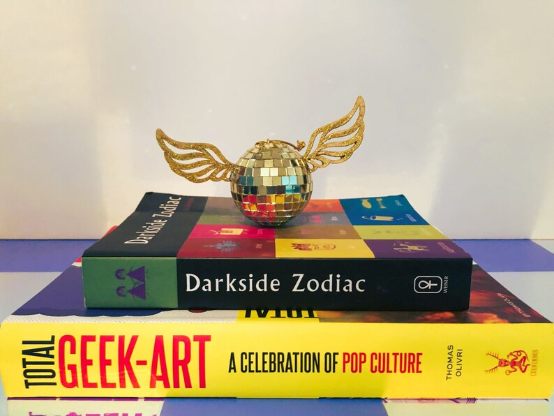 Puede incluir: Una bola de discoteca dorada con alas brillantes descansa sobre dos libros. El libro superior se titula "Darkside Zodiac" y el libro inferior se titula "Total Geek-Art: A Celebration of Pop Culture". Los libros est&aacute;n apilados sobre una superficie a rayas moradas y blancas.