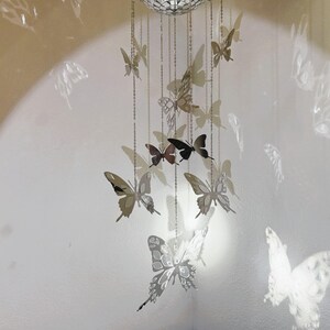 May include: A decorative mobile featuring multiple silver butterfly cutouts suspended from chains. The butterflies vary in size and are arranged in a cascading pattern. The background is a light-colored wall with a circular light.