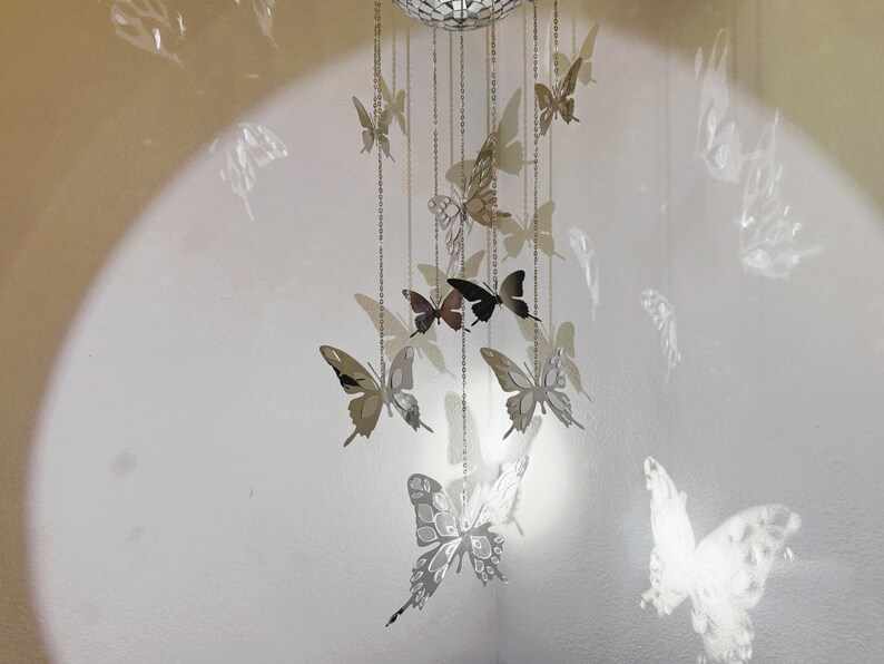 May include: A decorative mobile featuring multiple silver butterfly cutouts suspended from chains. The butterflies vary in size and are arranged in a cascading pattern. The background is a light-colored wall with a circular light.