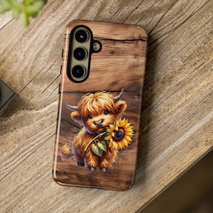 May include: A brown wooden phone case with a cute cartoon highland cow holding a sunflower. The case has a cutout for a triple camera lens.