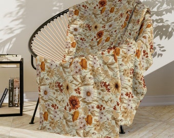 Könnte beinhalten: Eine floral gemusterte Decke, die über einem schwarzen Stuhl drapiert ist. Die Decke hat einen cremefarbenen Hintergrund mit einem sich wiederholenden Muster aus orangefarbenen, braunen und weißen Blumen und Blättern. Der Stuhl hat ein schwarzes Metallgestell und eine gewebte Rückenlehne.
