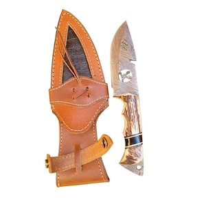 May include: A hunting knife with a patterned blade and a deer antler handle, next to a brown leather sheath. The blade has a decorative etching of a bird. The sheath has a strap and a decorative stitched design.