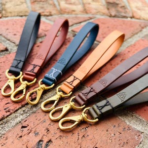 May include: Five leather wrist straps in various colors: black, brown, blue, orange, and olive green. Each strap features a gold-colored clasp and a stamped design. The straps are arranged on a brick surface.